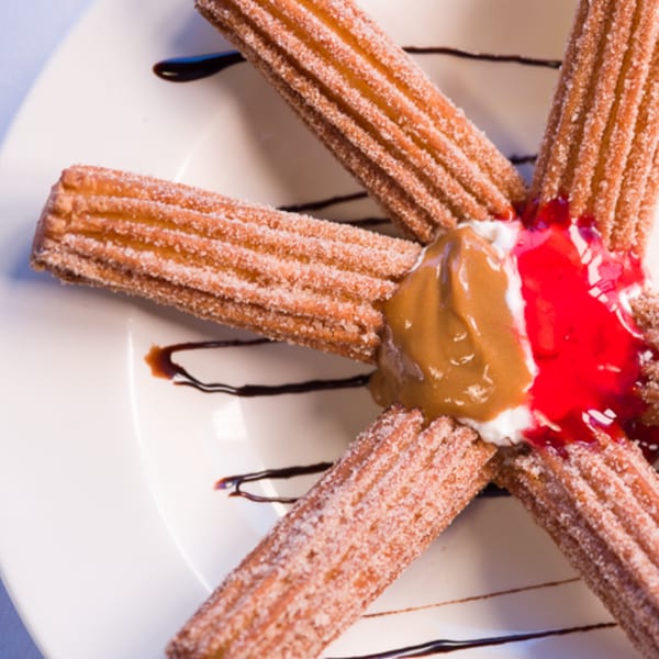 Five sugar-dusted churros arranged in a star, with caramel, whipped cream, and strawberry jam in the center.