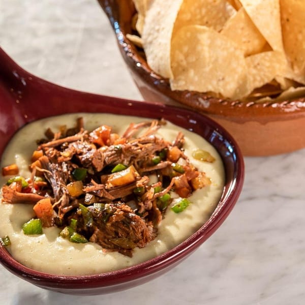 Creamy queso dip topped with shredded beef and diced vegetables, with a bowl of tortilla chips.